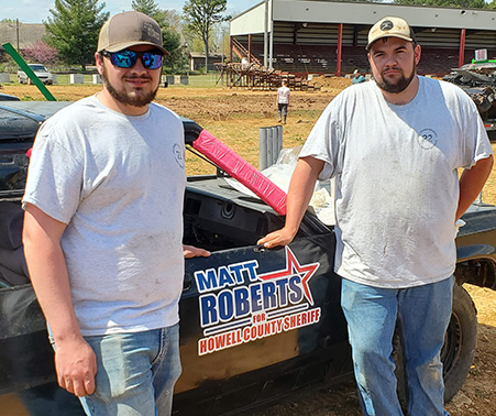 Dakota and Dustin Tiger standing in front of a demolition derby car displaying a Matt Roberts Logo